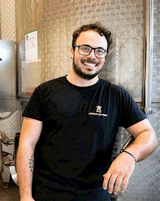 A person in a black shirt stands smiling in front of large metal equipment, possibly in an industrial or production setting.