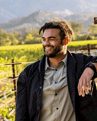 The image shows a person smiling outdoors in a vineyard with mountains in the background, enjoying a sunny day.