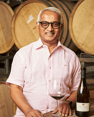 A person stands near wine barrels with a glass of red wine and a bottle on a wooden table, wearing a pink shirt and glasses.