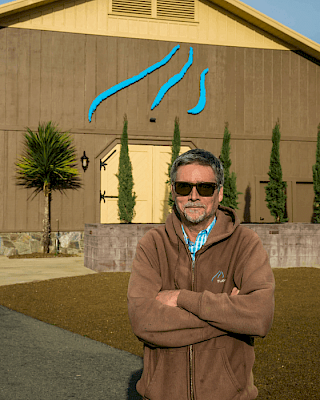 A person stands in front of a building with a blue logo, wearing sunglasses and a brown jacket.