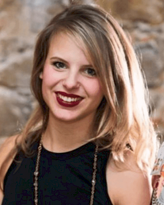 The image shows a person with shoulder-length hair, wearing a black top and a necklace, smiling at the camera.