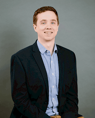 A person is sitting on a chair, wearing a blue shirt and dark blazer, with a neutral background.