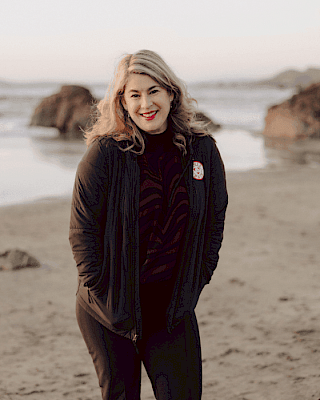 A person stands on a sandy beach with rocks in the background, wearing a dark outfit, smiling at the camera.