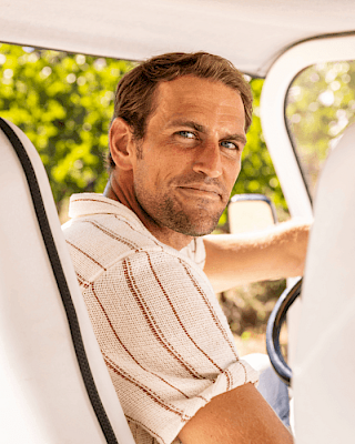 A man sitting in a vehicle, looking back over his shoulder, with greenery visible through the window.