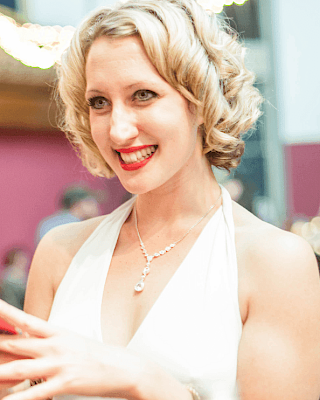 A woman in a white dress, smiling, with styled blonde hair and a necklace, stands at an indoor event with string lights in the background.