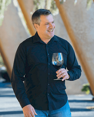 A person is smiling while holding a wine glass, standing outdoors with a background of wooden beams and greenery.