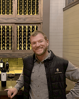 A person is standing in a wine cellar, smiling, with wine bottles and a glass on the counter.