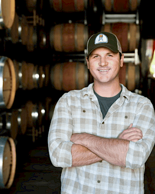 A person in a plaid shirt and cap stands smiling in front of rows of wooden barrels, possibly in a winery or brewery.