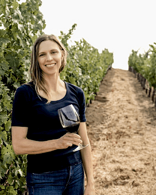 A woman stands in a vineyard holding a glass of red wine, surrounded by green grapevines under a clear sky.