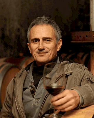 A person holding a glass of red wine, standing in a room with wooden barrels in the background, possibly a cellar or winery.