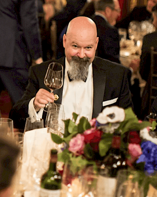 A man in formal attire holds a wine glass at an elegant dinner event, with floral centerpieces and other guests in the background.