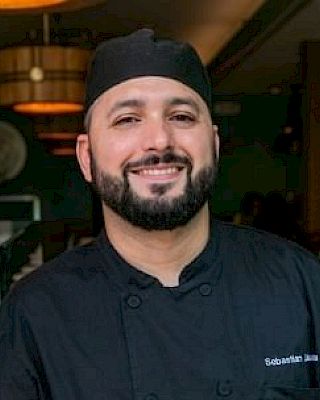 A smiling person wearing a black chef's uniform and hat, standing indoors.