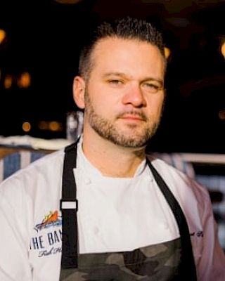 A person in a white chef's coat and black apron stands in a restaurant setting with blurred warm lighting in the background.