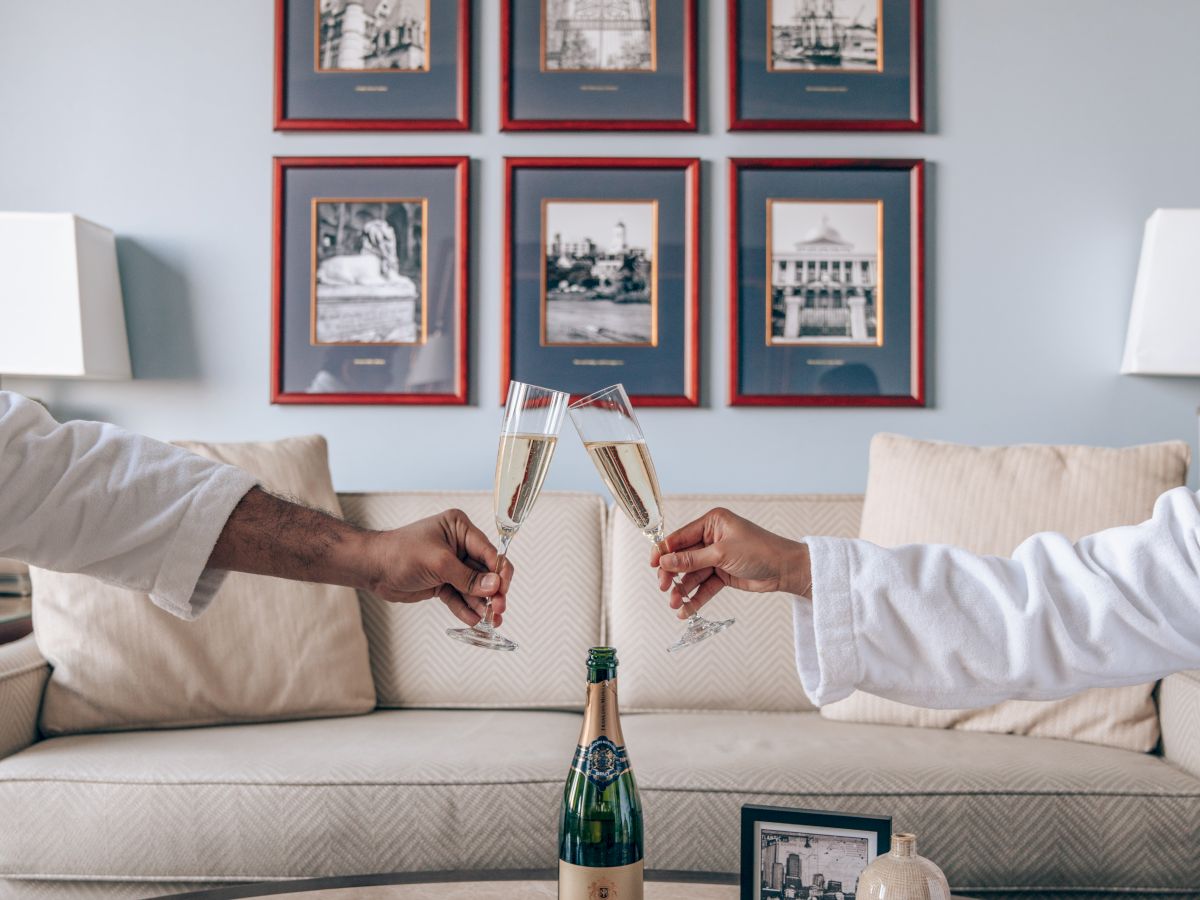 Two people in robes are toasting with champagne in a living room, with framed pictures on the wall and a coffee table in front.