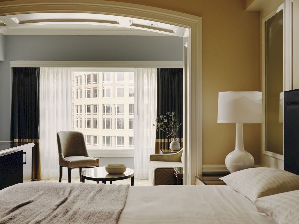 A cozy hotel room with a neatly made bed, a lamp, a chair, a small table, and large windows with cityscape views completes the scene.