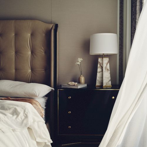 A bedroom scene with a tufted headboard, nightstand with lamp, vase, and book, and sheer curtains allowing light in.