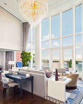 This image shows a luxurious, open-plan living and dining room with tall windows, elegant furnishings, chandeliers, and large potted plants.