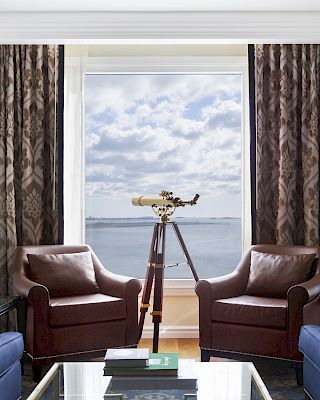 A living room with two brown chairs, a telescope by the window, blue sofas, and framed art on the walls. The view shows a body of water.