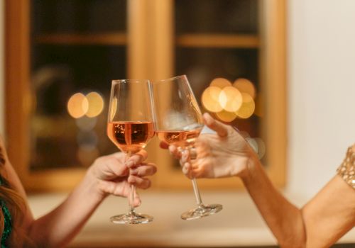 Two people are clinking wine glasses in a toast, with a softly blurred background of warm lights through a window.