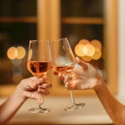 Two people are clinking wine glasses in a toast, with a softly blurred background of warm lights through a window.