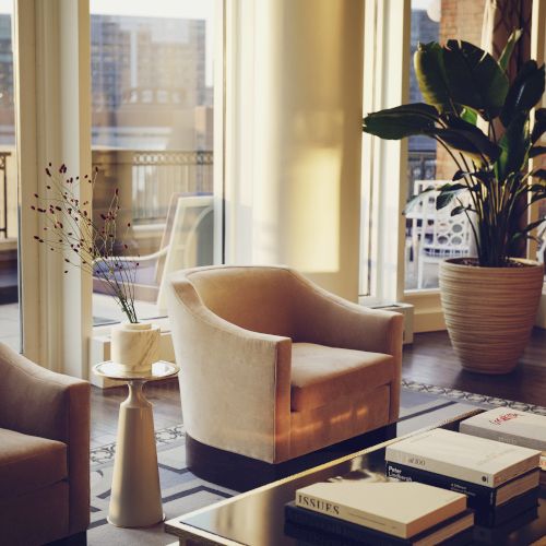 A modern living room with plush chairs, a potted plant, and a glass table topped with books, near large windows overlooking a city view.