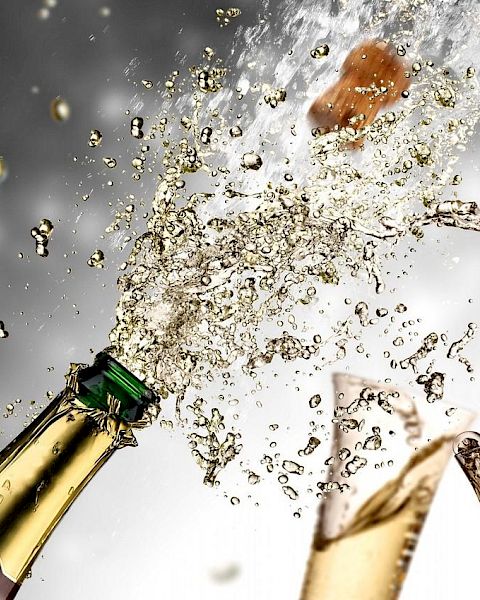 A champagne bottle being opened with liquid splashing and bubbles, alongside two filled glasses, creating a celebratory scene.