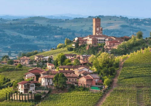 A picturesque hillside village surrounded by lush vineyards, with a prominent, tall tower and rolling green hills in the background.