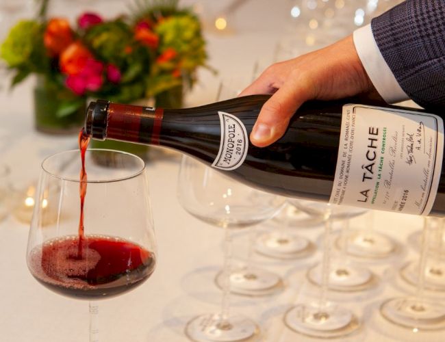 A bottle of La Tâche wine being poured into a glass at an elegant setting with flowers and neatly arranged empty glasses.