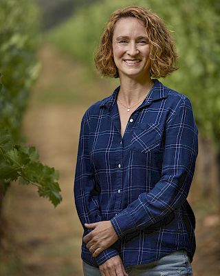 A person is standing in a vineyard, wearing a blue plaid shirt, smiling at the camera. The background shows rows of grapevines.