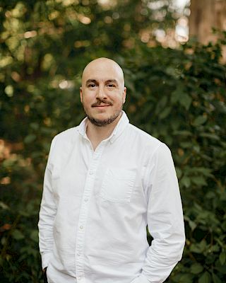 A person in a white shirt stands outdoors in front of lush, green foliage.