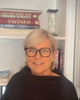 A person with glasses is smiling in front of a bookshelf with books and an hourglass.