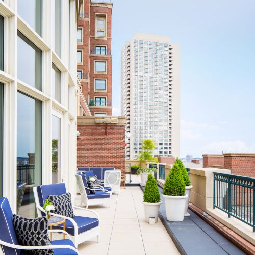 A rooftop terrace with blue and white chairs, potted plants, and views of city skyscrapers under a clear sky.