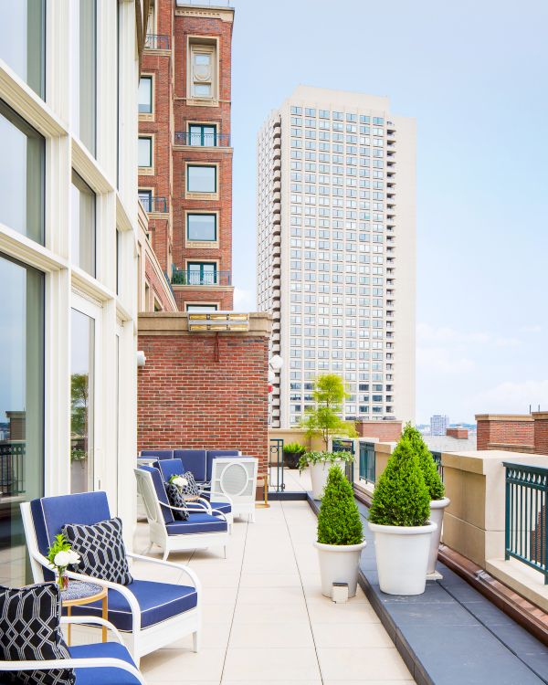 A rooftop terrace with blue and white chairs, potted plants, and views of city skyscrapers under a clear sky.