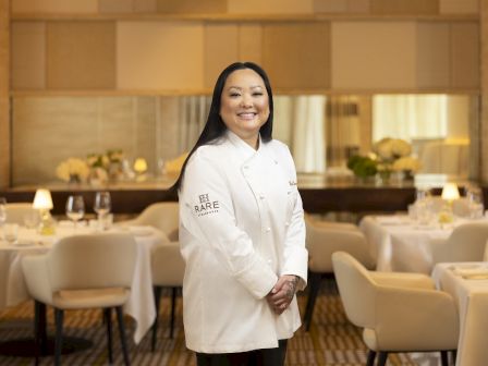 A chef in a white coat smiles in an elegant dining room with neatly arranged tables and chairs, featuring subtle decorative elements.