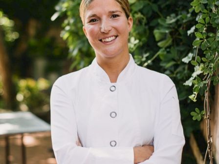 A person in a white chef's coat stands outdoors, smiling with arms crossed, next to greenery and a table.