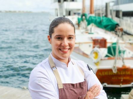 A person in a chef's uniform stands smiling by a waterfront with boats in the background.